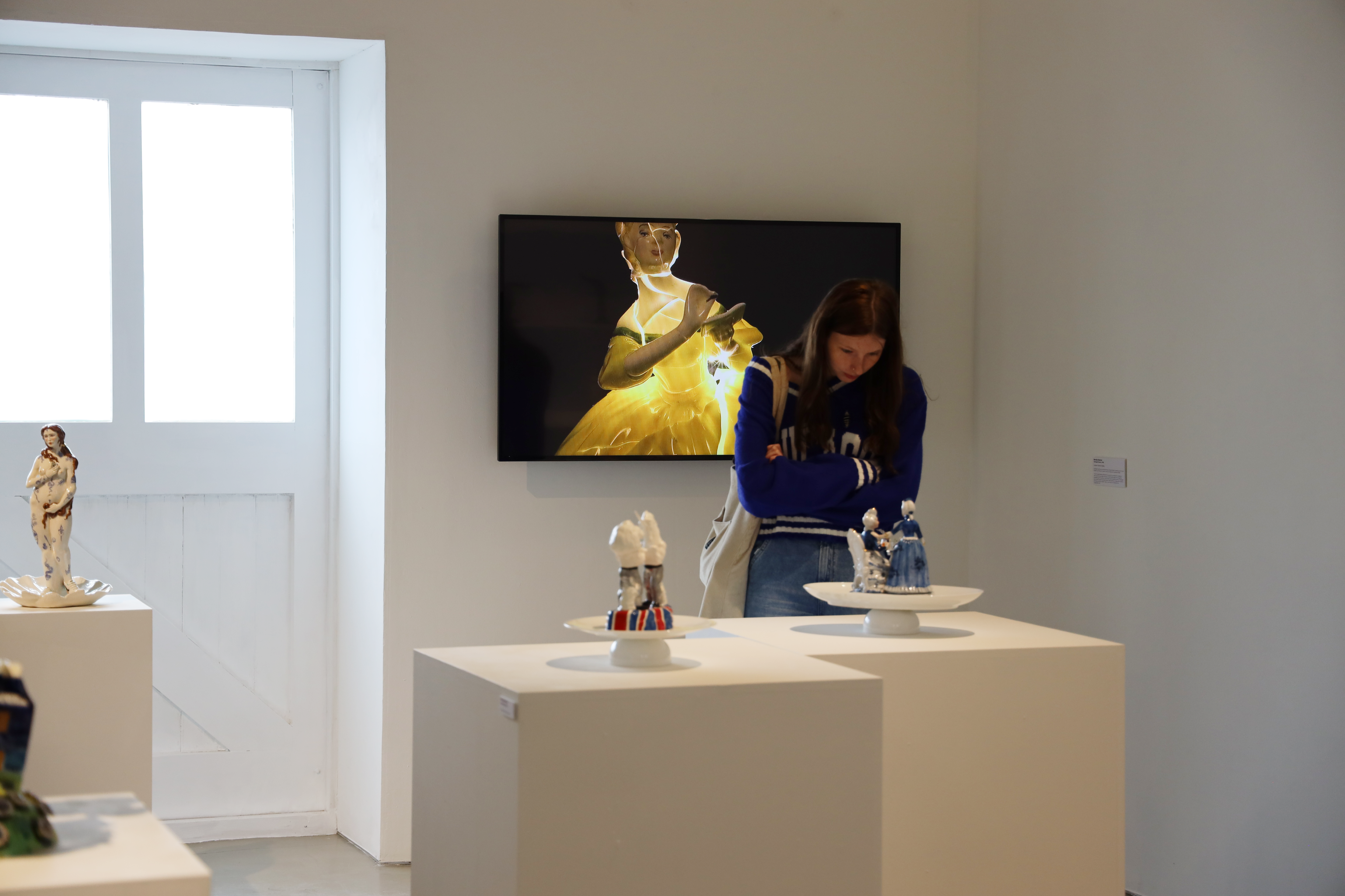 A student looks at a ceramic sculpture in a gallery with a large image of a broken, but mended ceramics figure behind her.