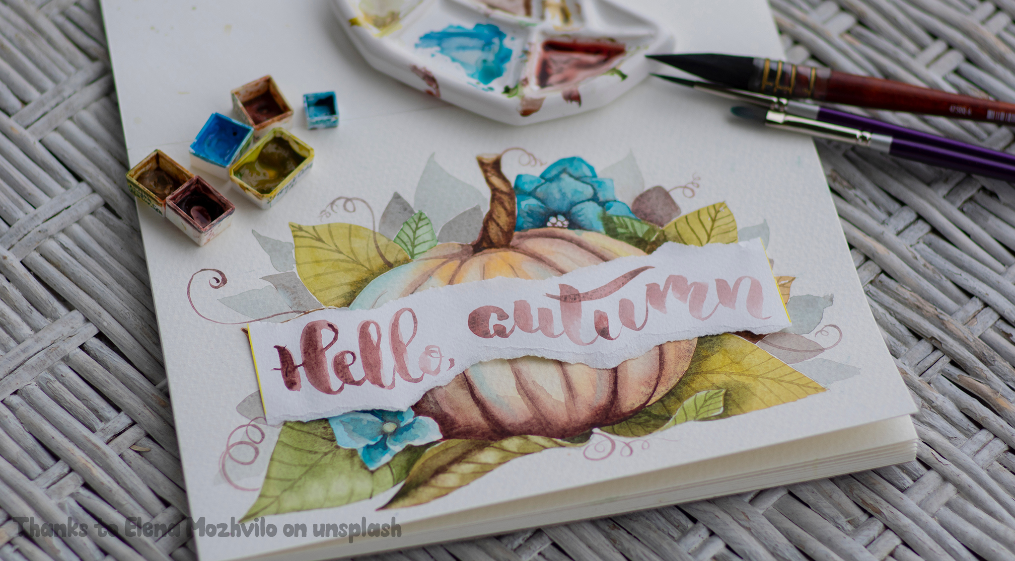 A beautiful painting of a watercolour pumpkin with hand written calligraphy which reads 'hello autumn' the pumpkin is a warm brown, there is blue flowers and green leaves and curling vines in the background. Brushes and watercolour paint squares are above the illustration