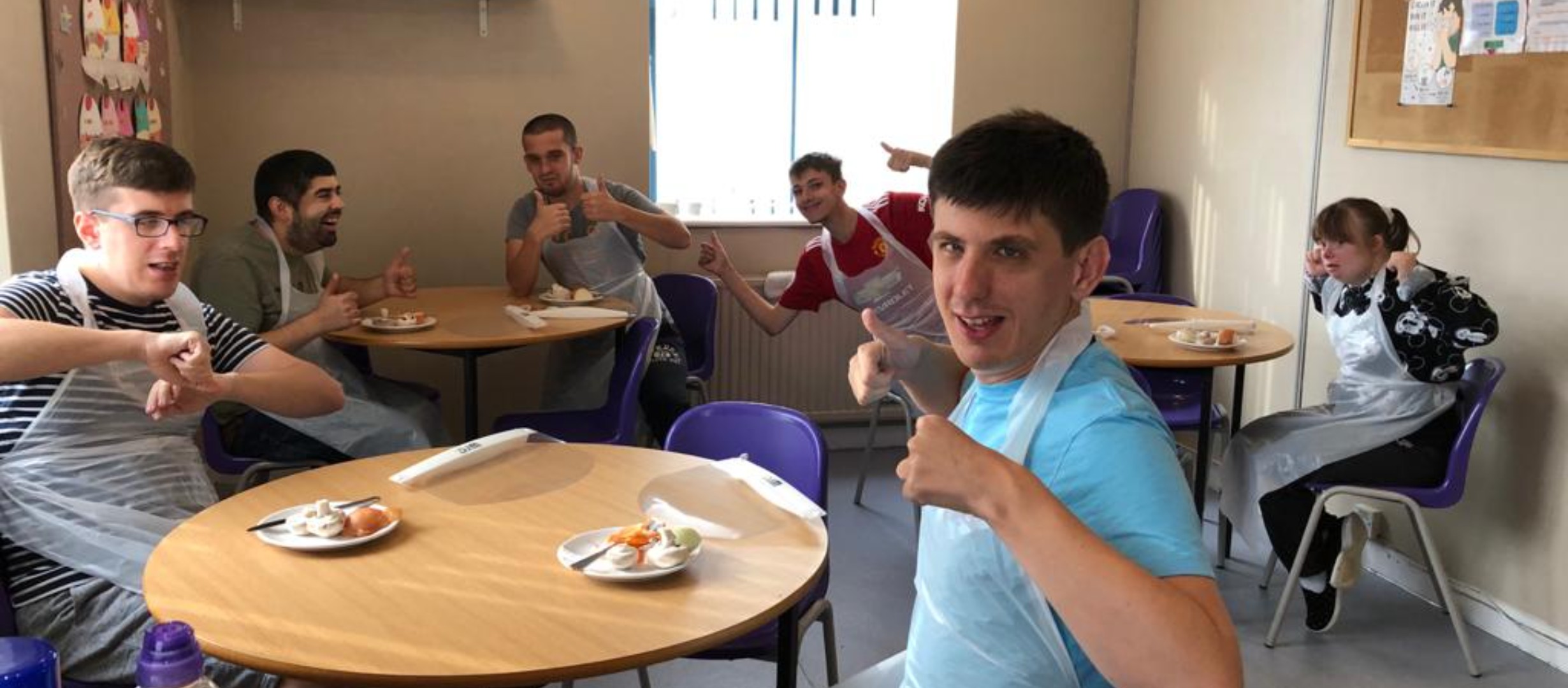young adults sitting at tables holding their thumbs up to camera with their lunch on the tables