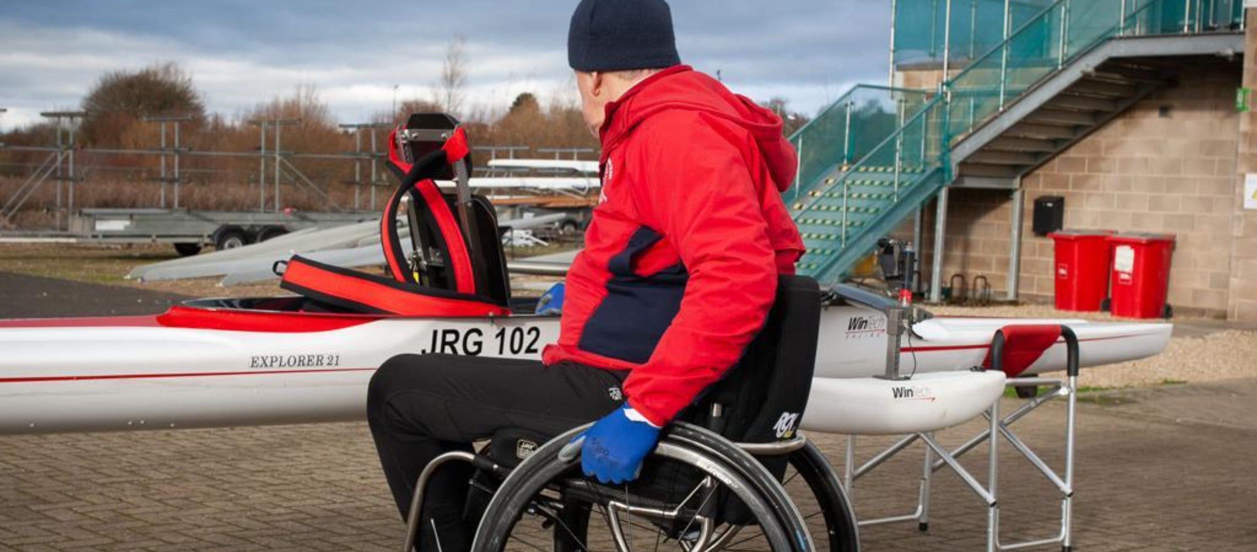 Rowing Boat on land with a rower in a red coat looking at it in his wheelchair. He is about to go on the water and is checking his equipment