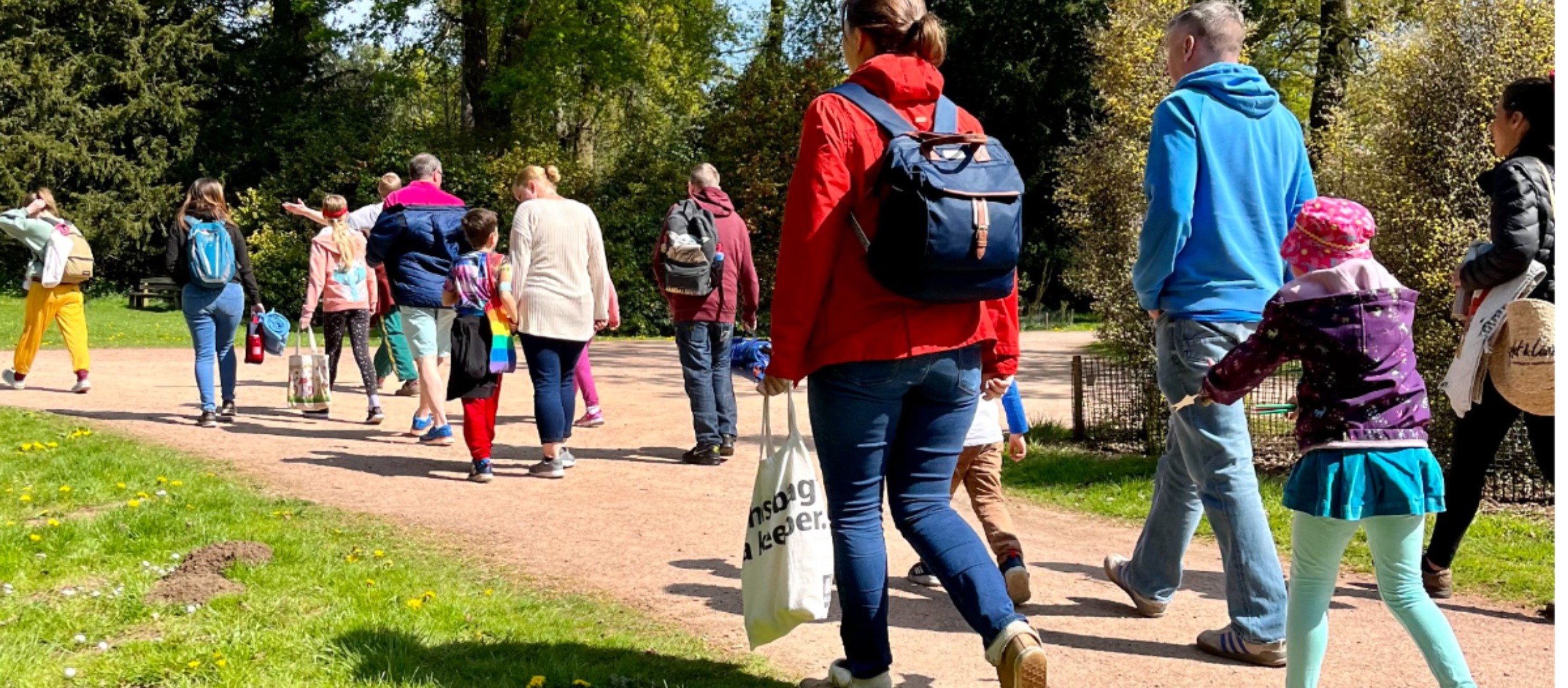 A colour photo of a group of families walking on a gravel path at Westonbirt Arboretum. They are surrounded by grassy fields, tall green trees and blue sky.