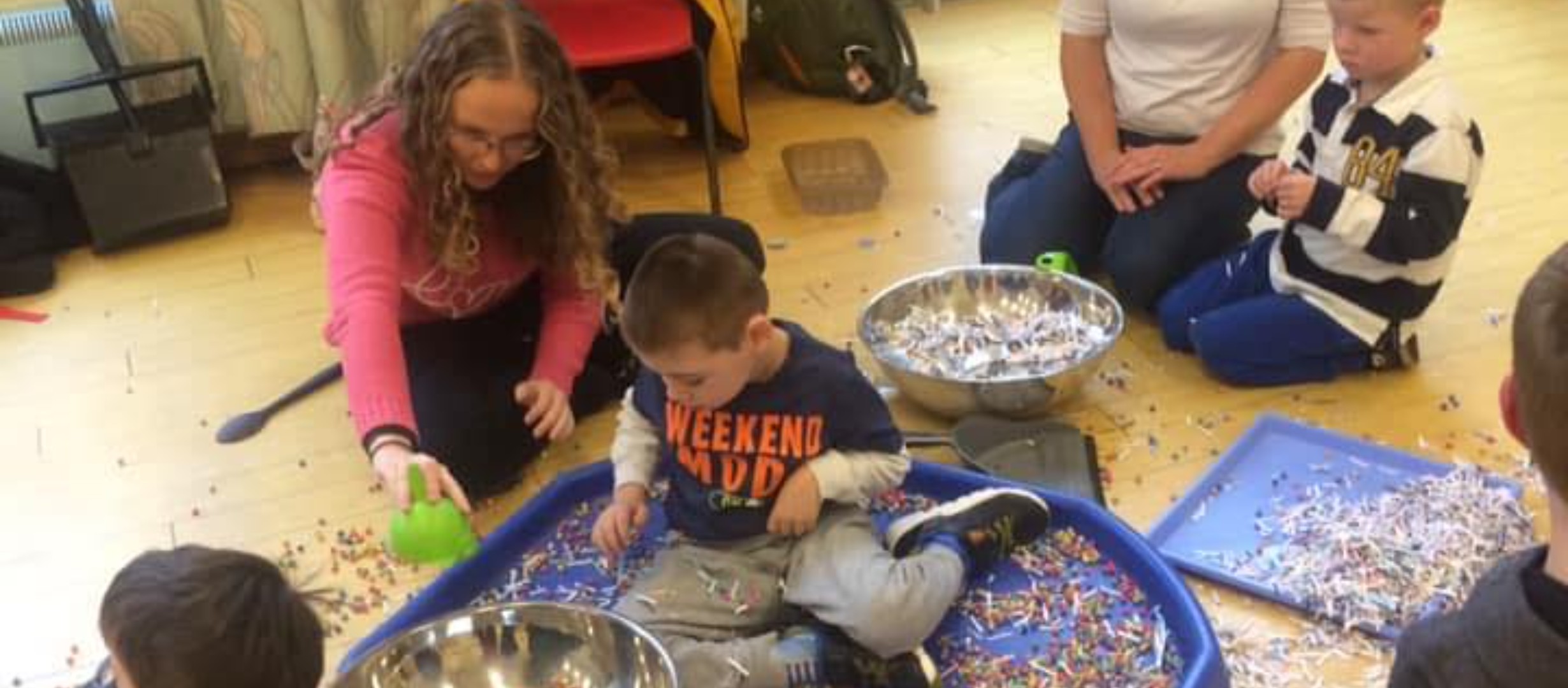 Boy in tuff tray with coloured chick peas, lady holding out an item