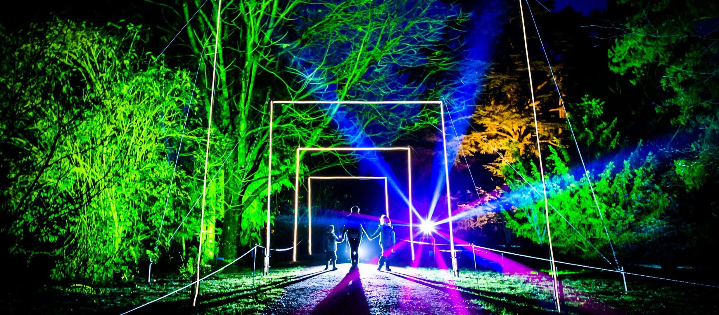 A dark night is brightened by a landscape of trees brightly lit up in greens, oranges and blue. A woman and her two young children stand between a square arch of light. A bright spot light shines from behind them.