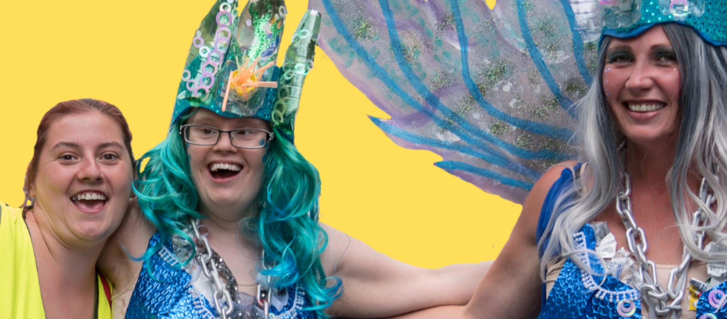 Two individuals smiling at the camera. Two are dressed in blue carnival outfits including crowns and a wing structure. The middle individual has blue curly long hair. The third person is smiling at the camera wearing a yellow top.