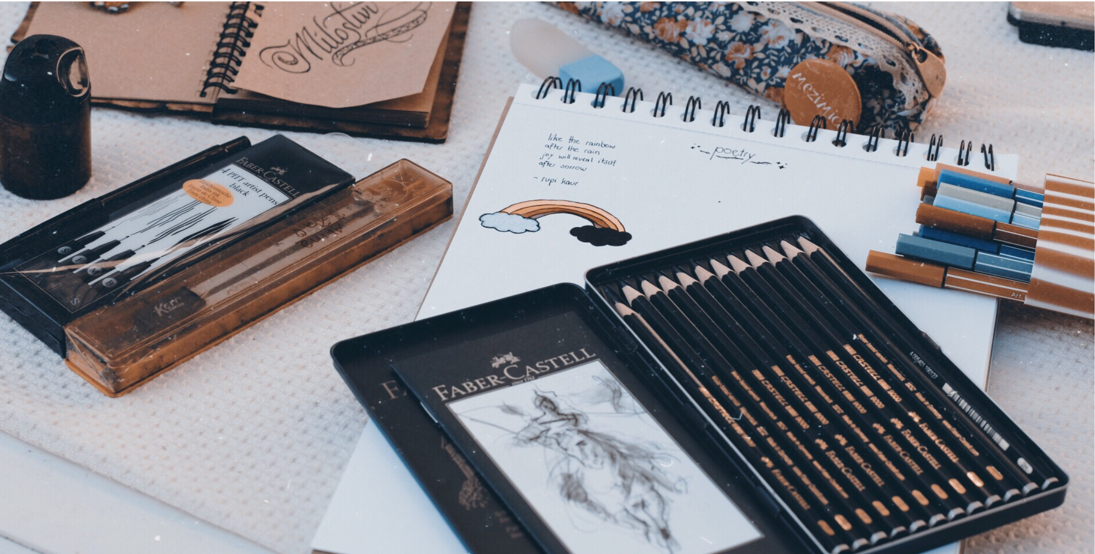 A photograph of a sketchbook with white pages and a sketchbook with brown pages. There are pencils and pens displayed around it and small sketches on the pages