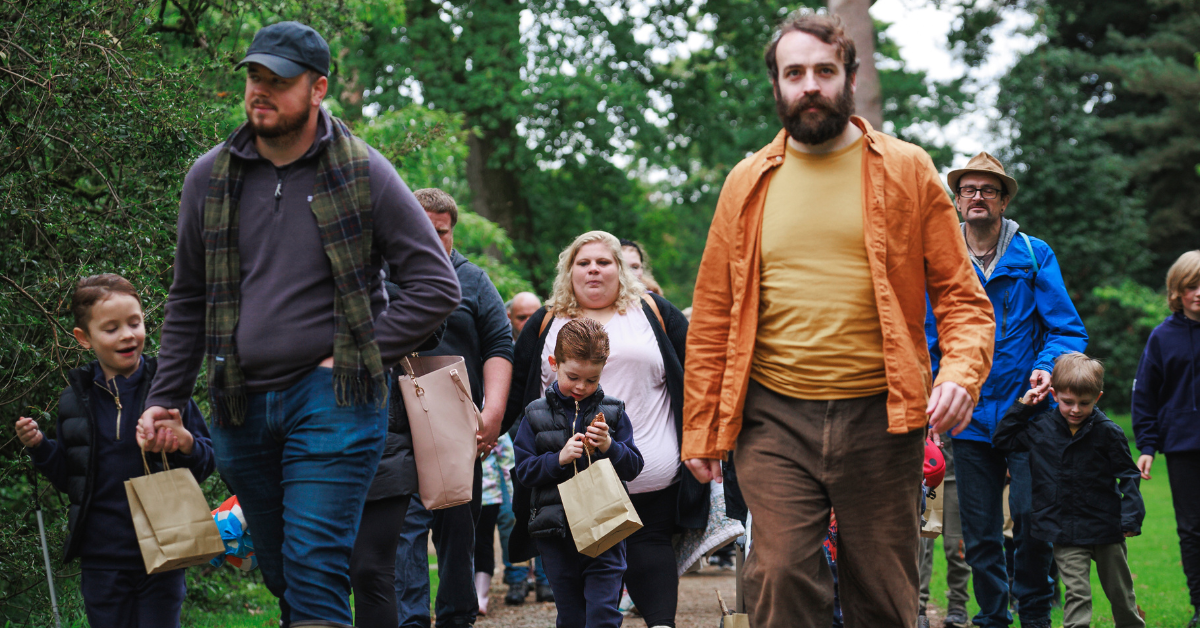 Colour photo of a group of families walking along a gravel path at Westonbirt Arboretum, with tall green trees standing behind them.