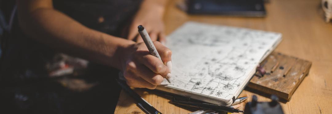 The photograph is is of a woman drawing in a sketchbook on a wooden table. She is holding a fine liner pen and has other pencils and pens spread around her. She is cast in a warm glow