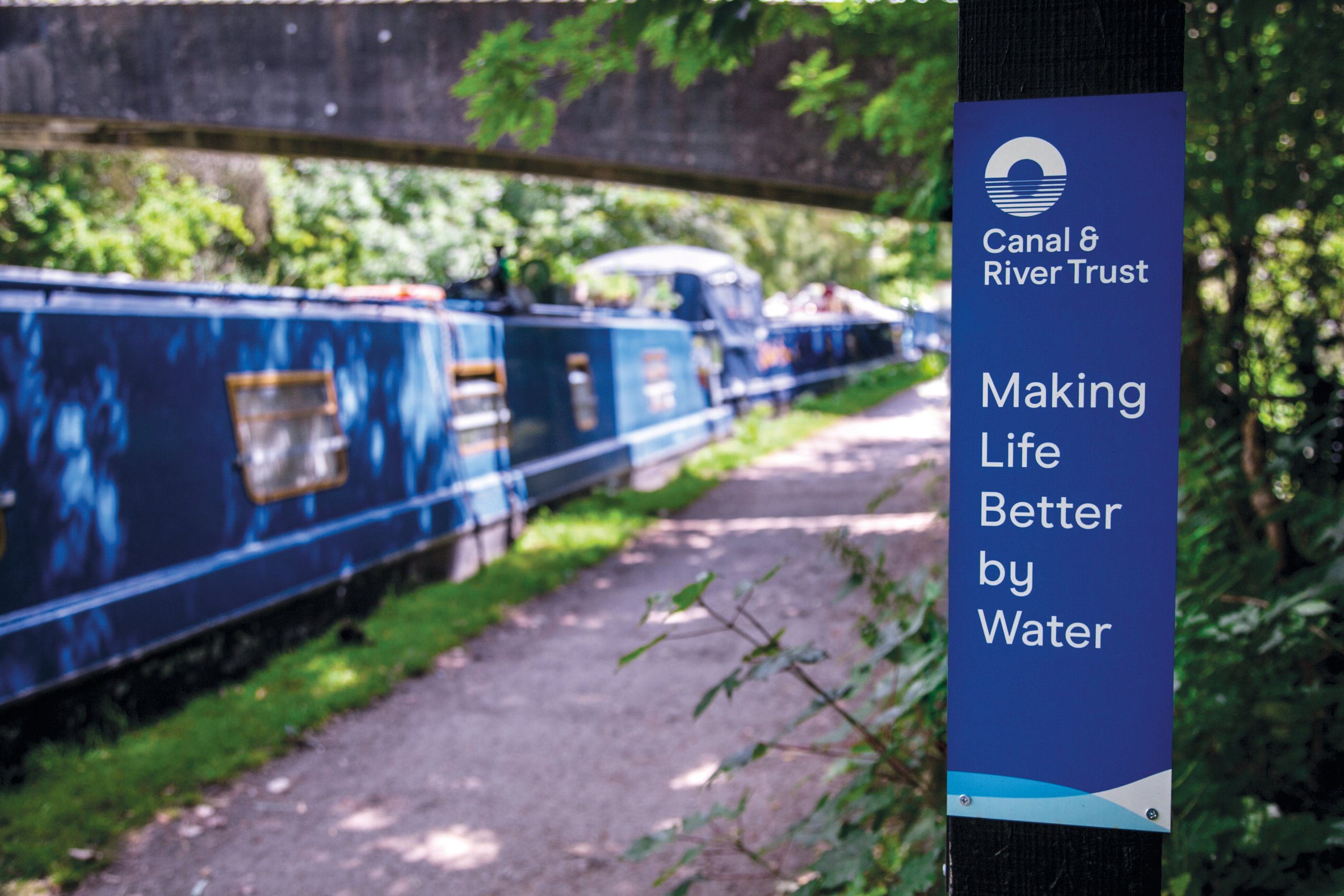 A blue canal boat moored alongside a paved towpath. A sign says 'Making life better by water' and show the Canal and River Trust Logo.