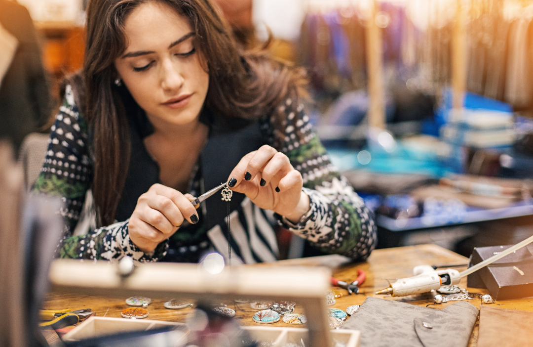 A young woman with dark hair is seated at a table. She is carefully crafting some jewellery using a tool to attach a charm to some thread
