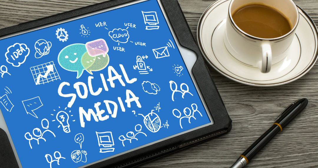 This photograph is of a tablet with a vibrant blue screen and the words SOCIAL MEDIA in large letters in the centre. Around it are white icons which relate to social media posts. The tablet is on a table with a pen and a cup of coffee