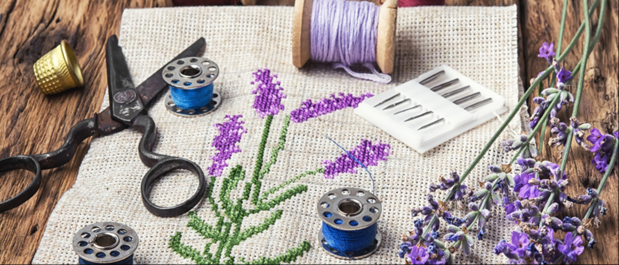 This is a photograph of a cross stitch with a lavender theme. There is lavender thread, scissors, and sprigs of lavender