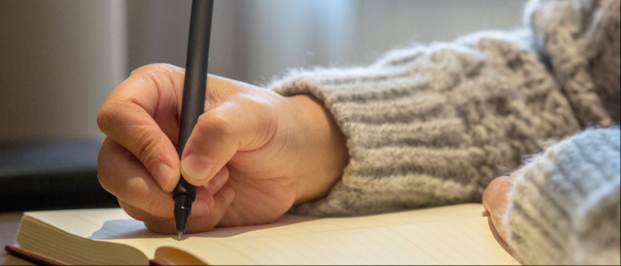 This is a photograph of someone writing in a notebook using a black pen. They are wearing a grey jumper