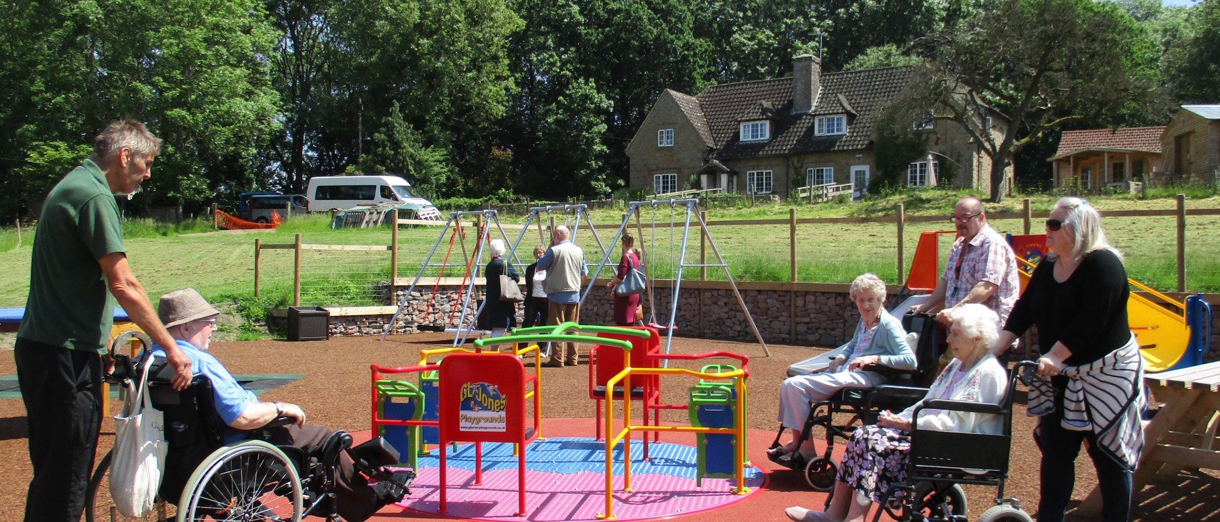 Image shows the Orchard Trust activity park with three people in wheelchairs and their carers about to use the roundabout. The Orchard Trust Orchards Care Home is in the background