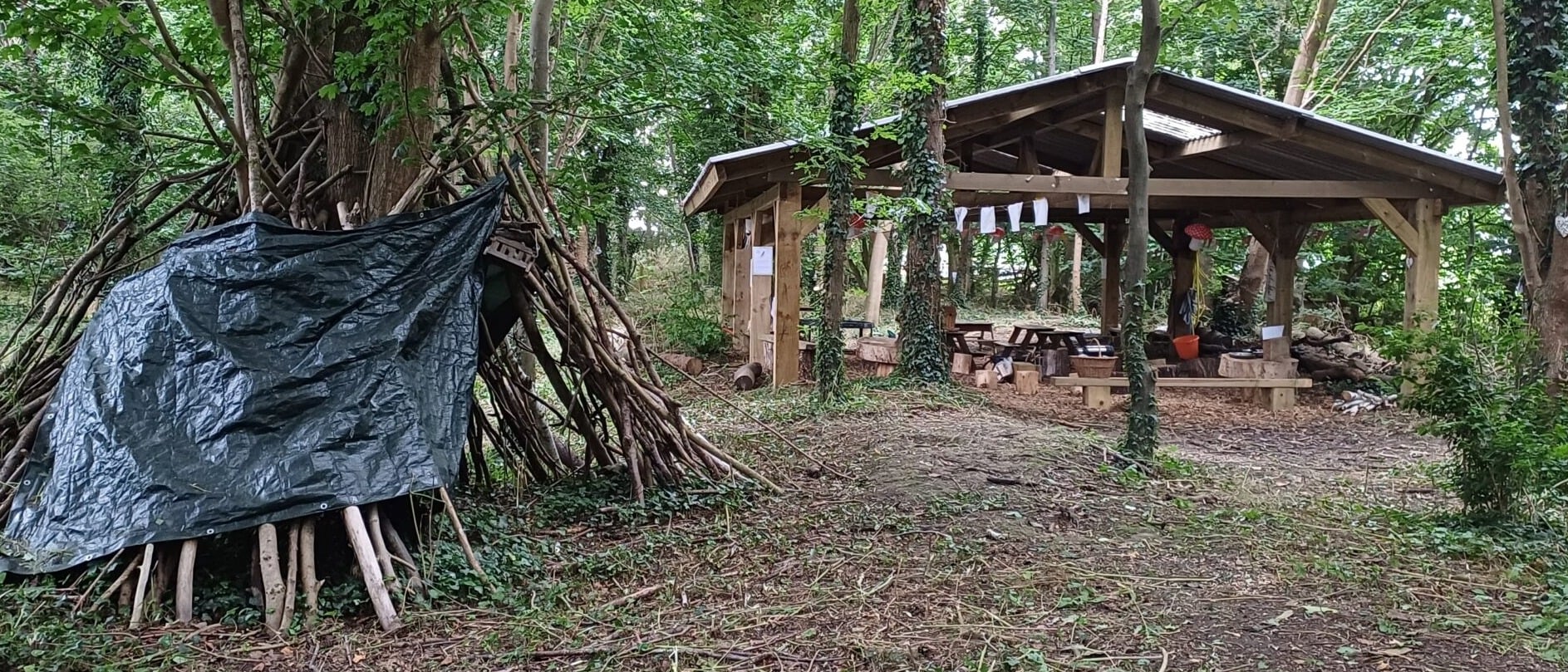 A woodland outdoor learning area with a wooden shelter on the right, furnished with benches and tables under a pitched roof. The shelter is decorated with paper signs and hanging items. On the left, a den made of sticks and a black tarp is built around a tree. The area is surrounded by dense green trees and natural forest ground.