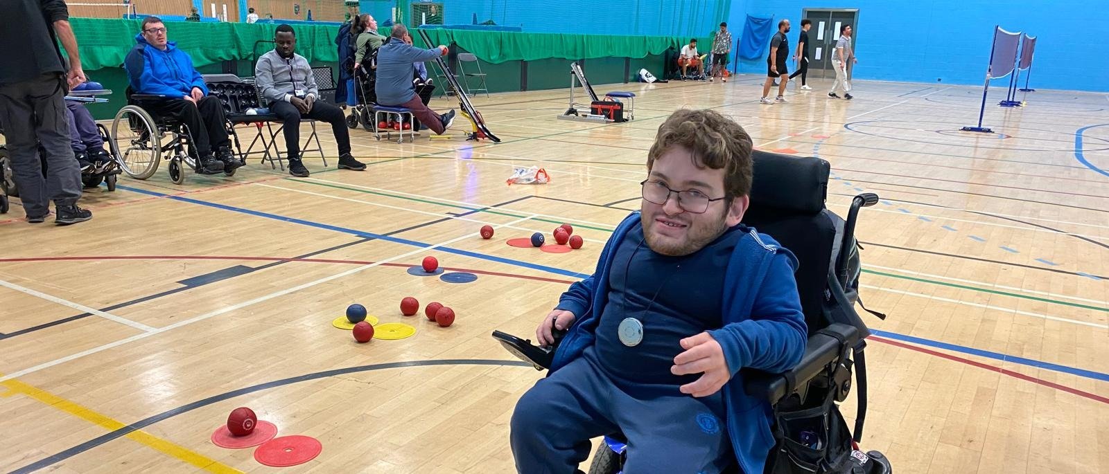 In the foreground at the right hand side you can see Archie, our boccia coach, who is sitting in a wheelchair. behind him are boccia balls our on court with a row of players sitting at the back in the far left of the image