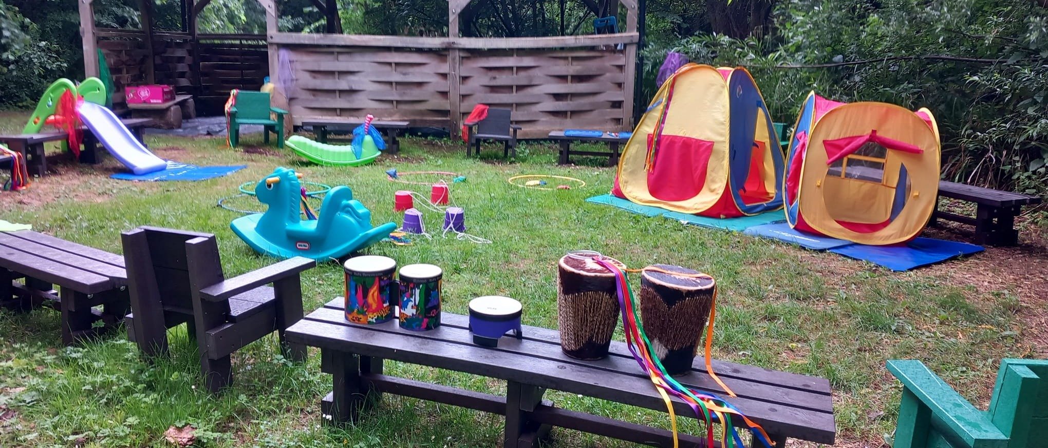 An outdoor wooden classroom and outdoor benches with colourful children's tents, play equipment and musical instruments, set within a woodland.