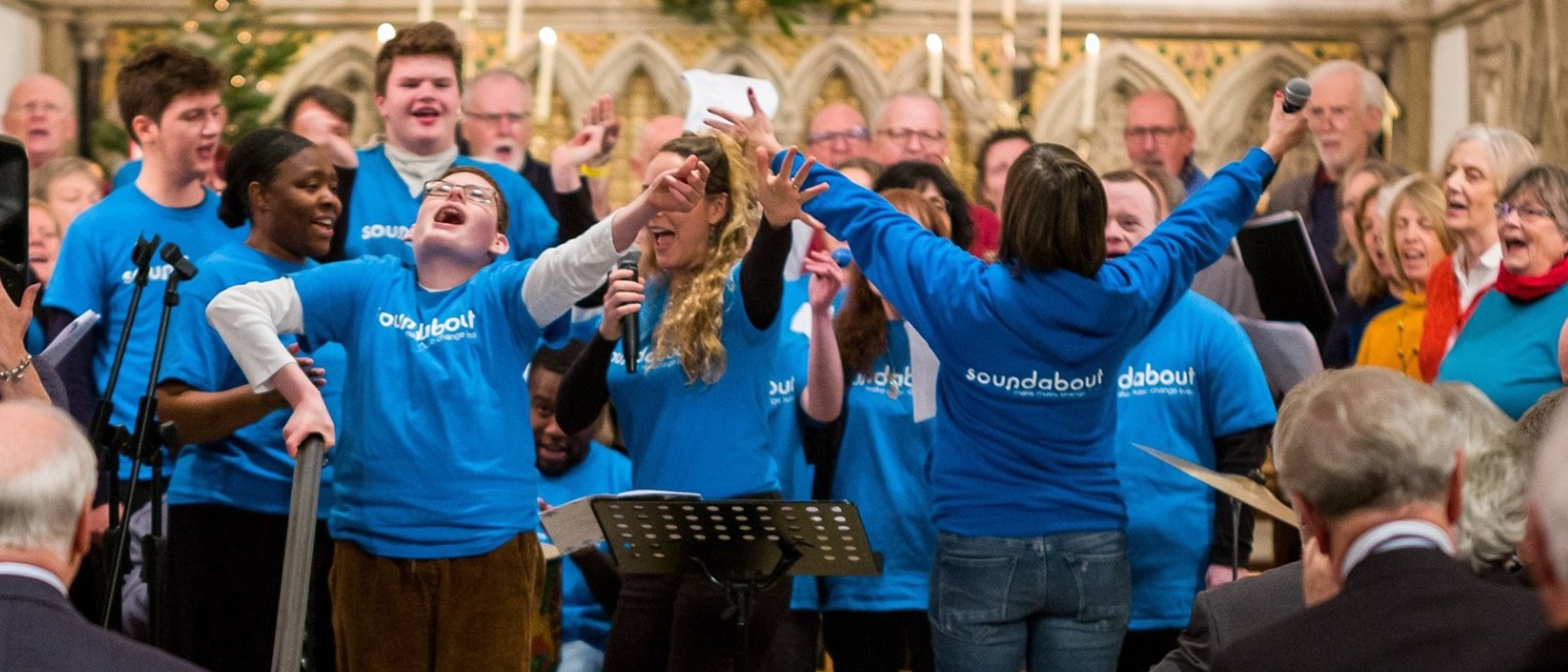 choir members performing. They all wear blue t-shirts and are singing or playing musical instruments